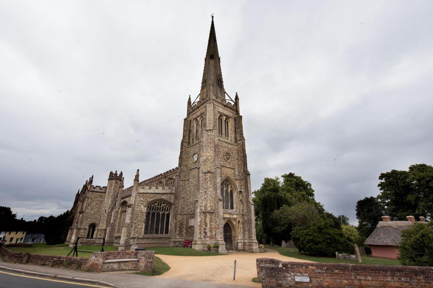 A medieval church and spire against a cloudy sky, surrounded by a lawn and driveway area. In the foreground is a road with a single yellow line.
