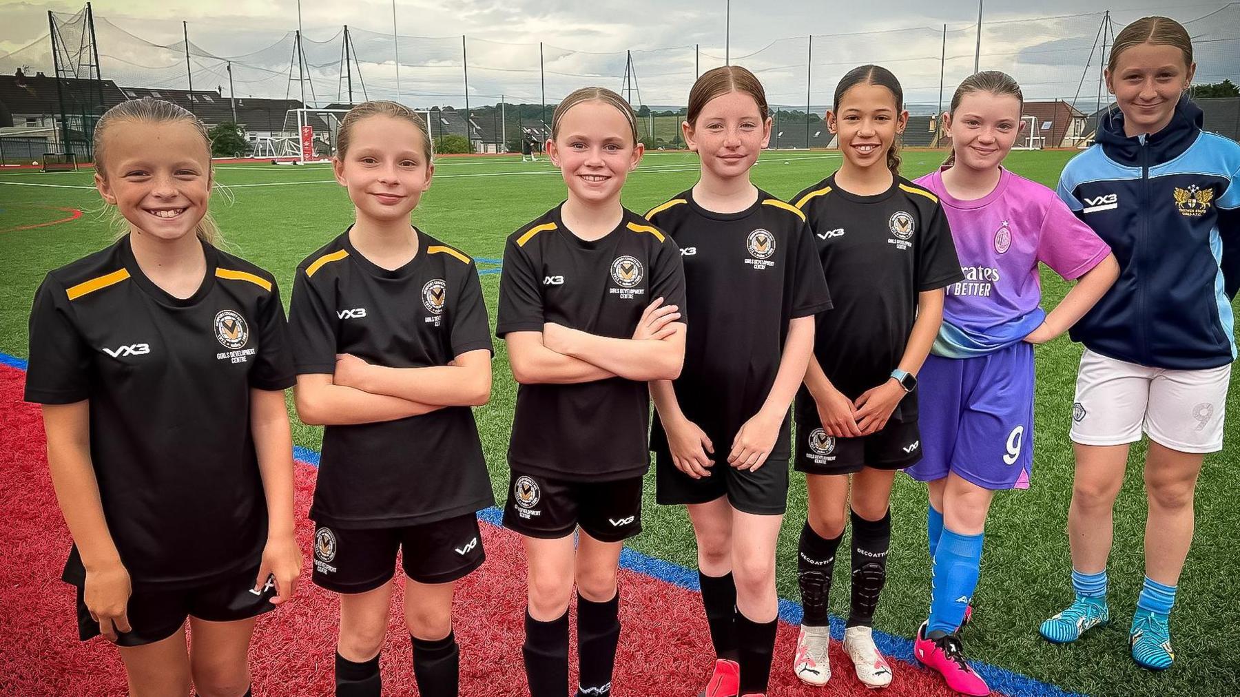 Girls standing together pitch side wearing their black Newport Country strip with gold strips on their shoulders