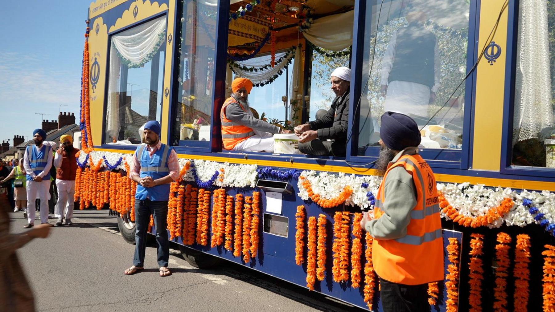 A yellow and blue vehicle is adorned with orange and white flowers. Two men one wearing an orange turban the other white are sitting at an open windo on the float. Other men wearing high vis jackets stand beside the vehicle. 