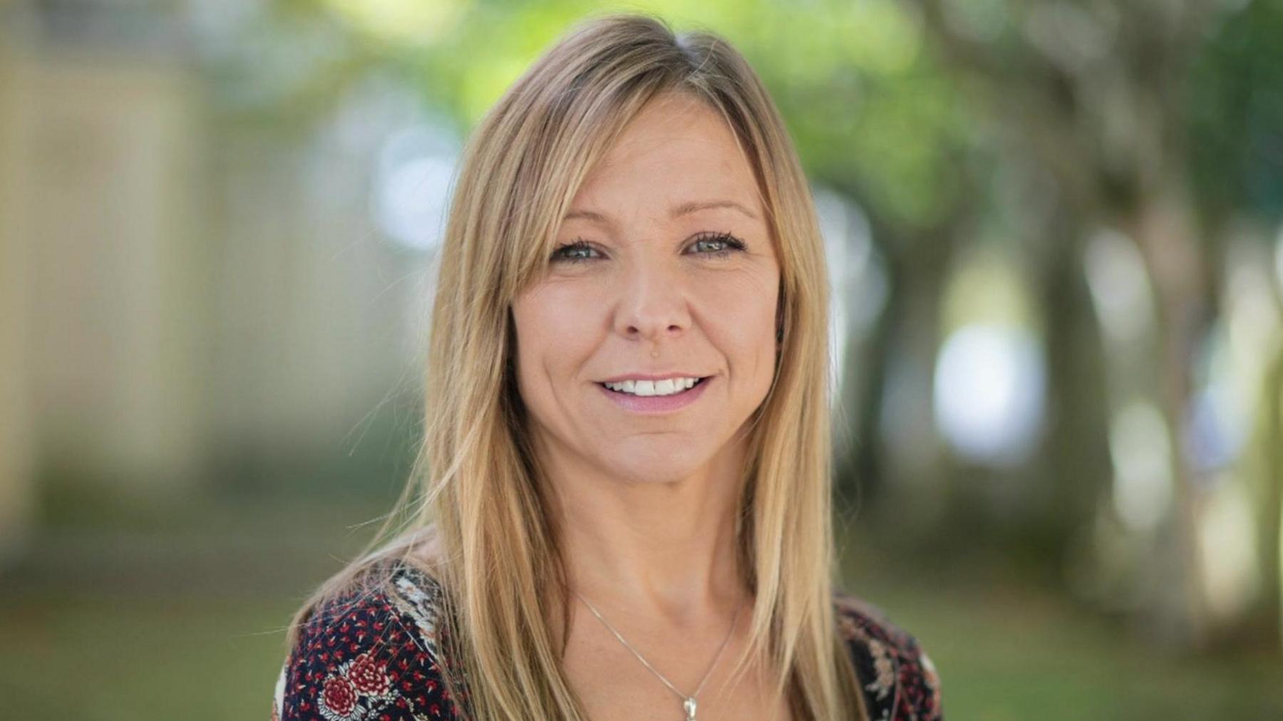 Claire Chick, who is smiling and has straight, shoulder-length hair. She is in an outdoor setting and is wearing a patterned top with a subtle necklace.