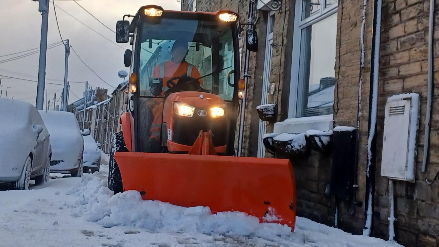 A man driving a bright orange gritter drives down a road covered in snow. Cars parked nearby are completely covered in snow.