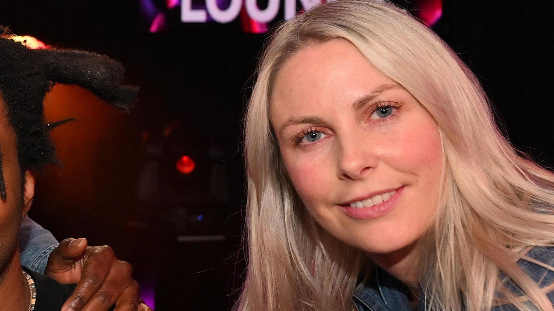 Charlie Hedges with long blonde hair smiling at the camera against a dark background. The bottom of the word "Lounge" in white letters is visible behind her.