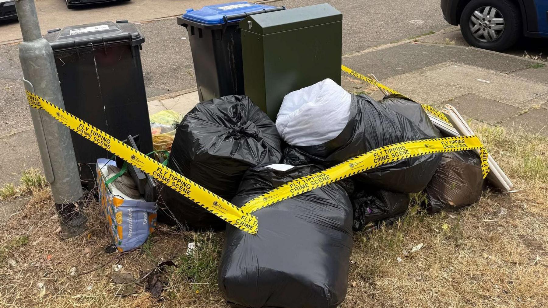 A large pile of rubbish, with tape round it, next to two wheelie bins and a green metal cabinet. The bin bags and bits of rubbish are on a grass area, with a car to the right.