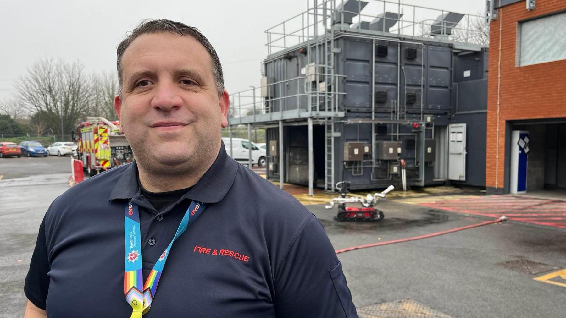 A man in a blue polo shirt stands in front of Ramsgate fire station - the fire robot is in the forecourt behind him.