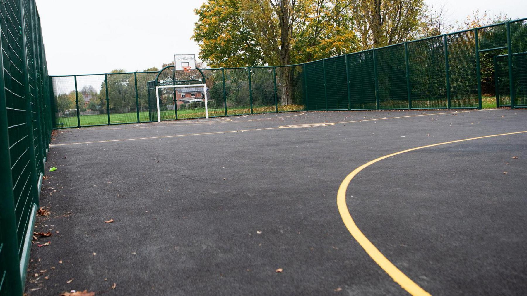 A multi-use games area with a grey surface, yellow lines and a football goal with a basketball net above at the end. The area is enclosed by green wire fencing.