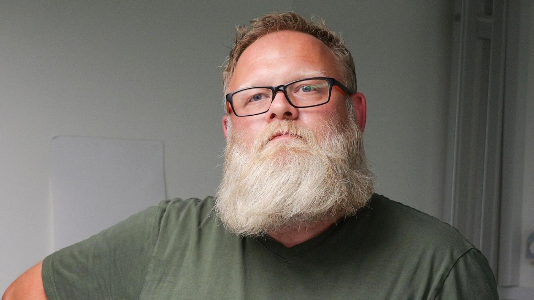 Aaron Clark is a bearded man in glasses. He is stood in a hospital ward.