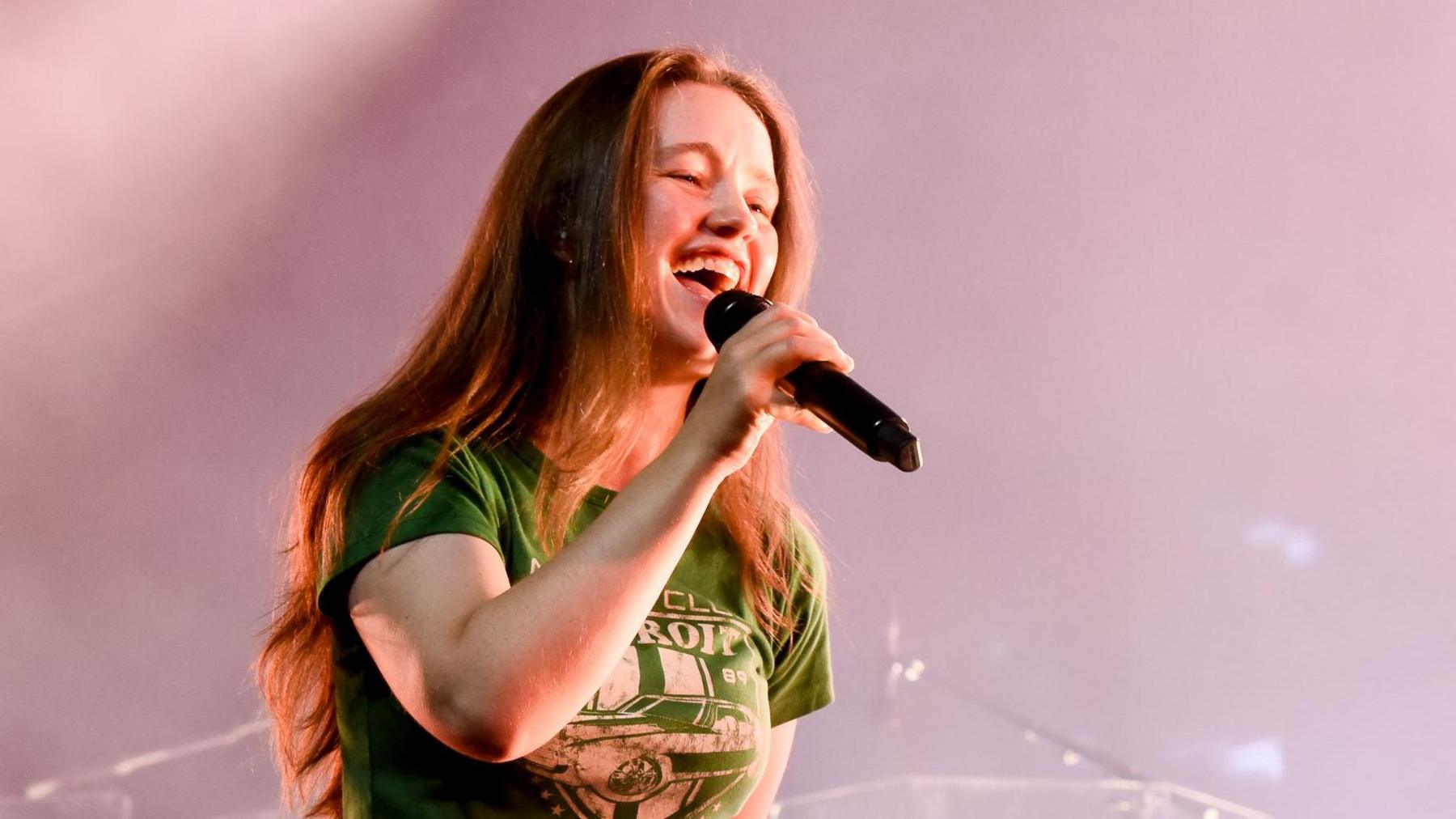 Sigrid is singing into a mic smiling. She has long light brown hair, a green t-shirt on and denim jeans. Behind her on the stage is a blond woman playing the bass guitar. There is also a drum set behind her.