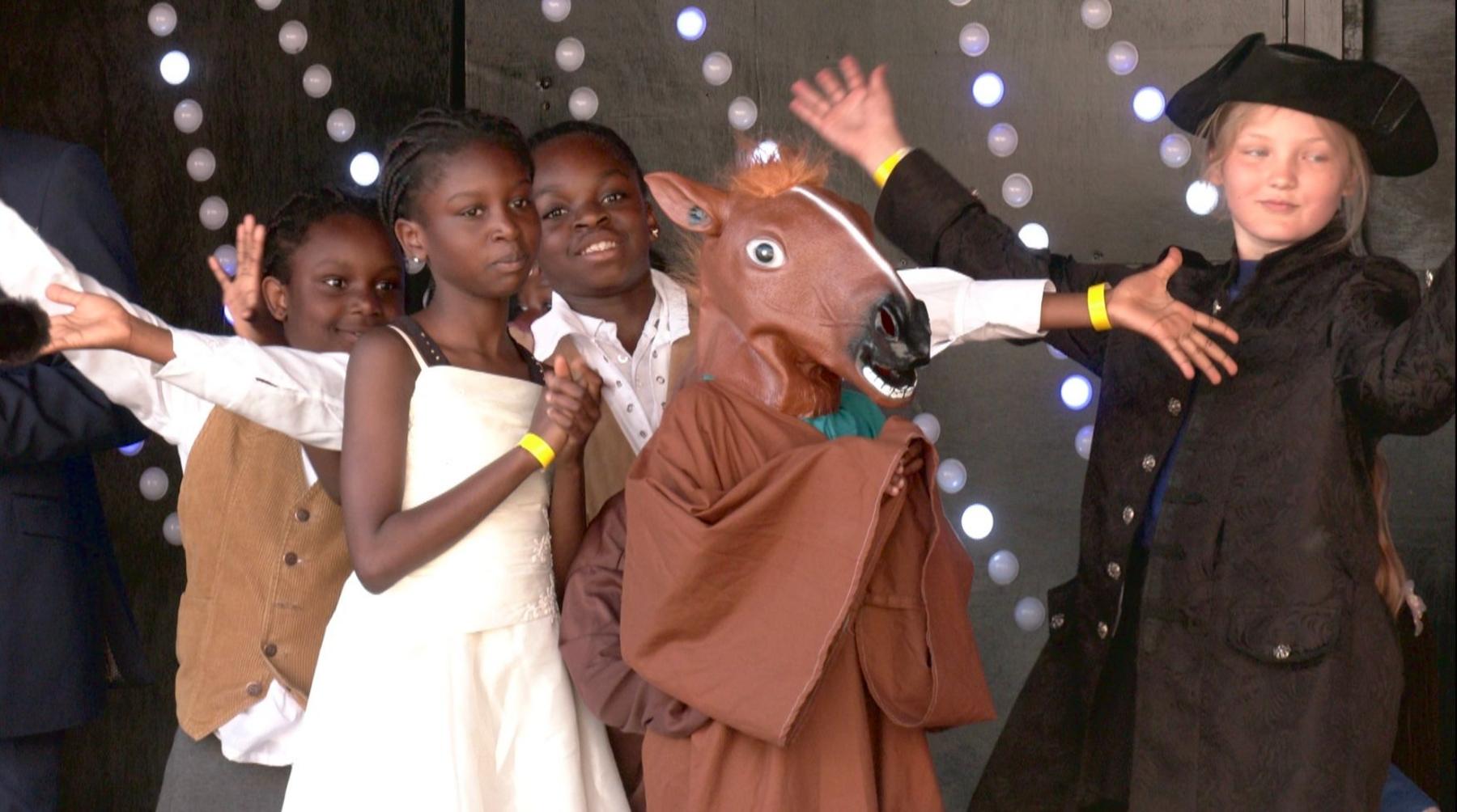Four children on stage performing. Two boys and two girls. One girl is at the front in a white dress, her hands are clasped together. The three behind her have their arms wide open and another person is dressed in a brown horse costume.