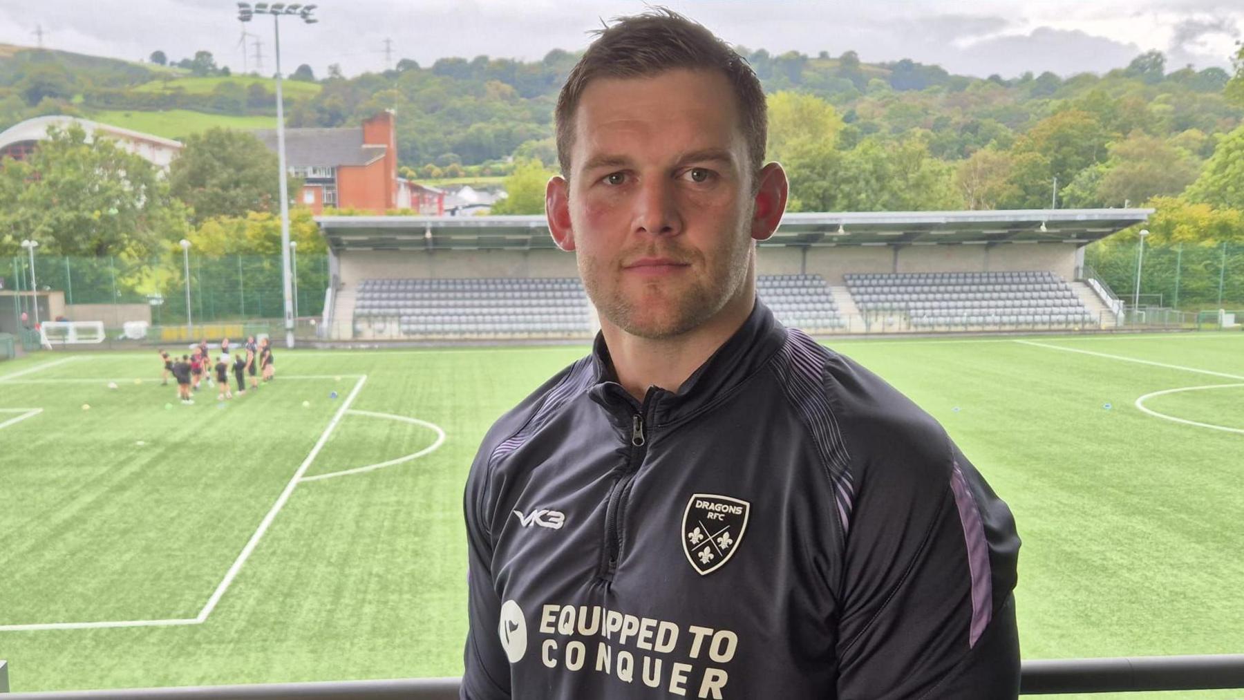 Dan Lydiate wearing his black kit, standing in front of a rugby pitch