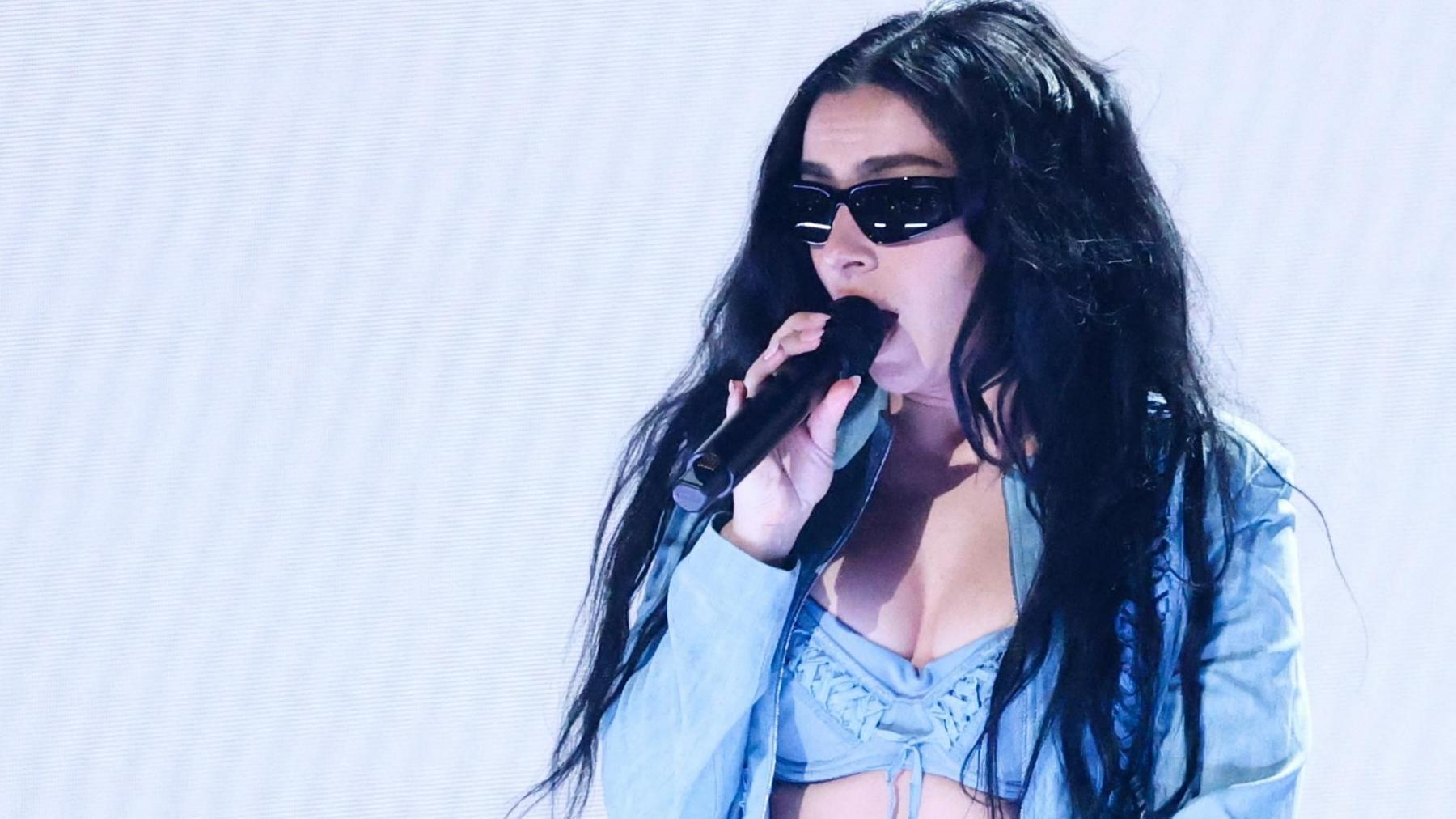 Charli XCX in an open blue top with blue bra and bare flesh visible. She is wearing sunglasses and singing into a hand-held microphone.