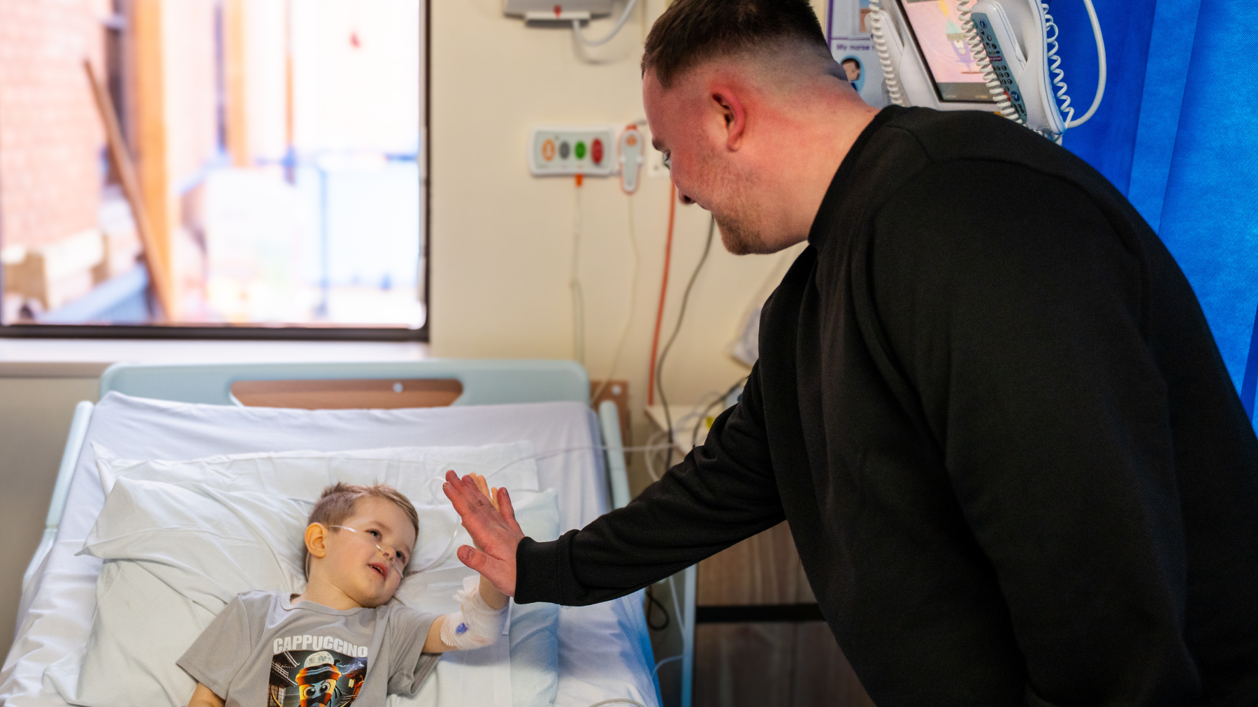 A child lies in a hospital bed with medical equipment nearby, wearing a grey T-shirt with a graphic print. Luke wears a black sweatshirt and leans forward to give the child a high-five. The room has a blue privacy curtain and a window letting in natural light.