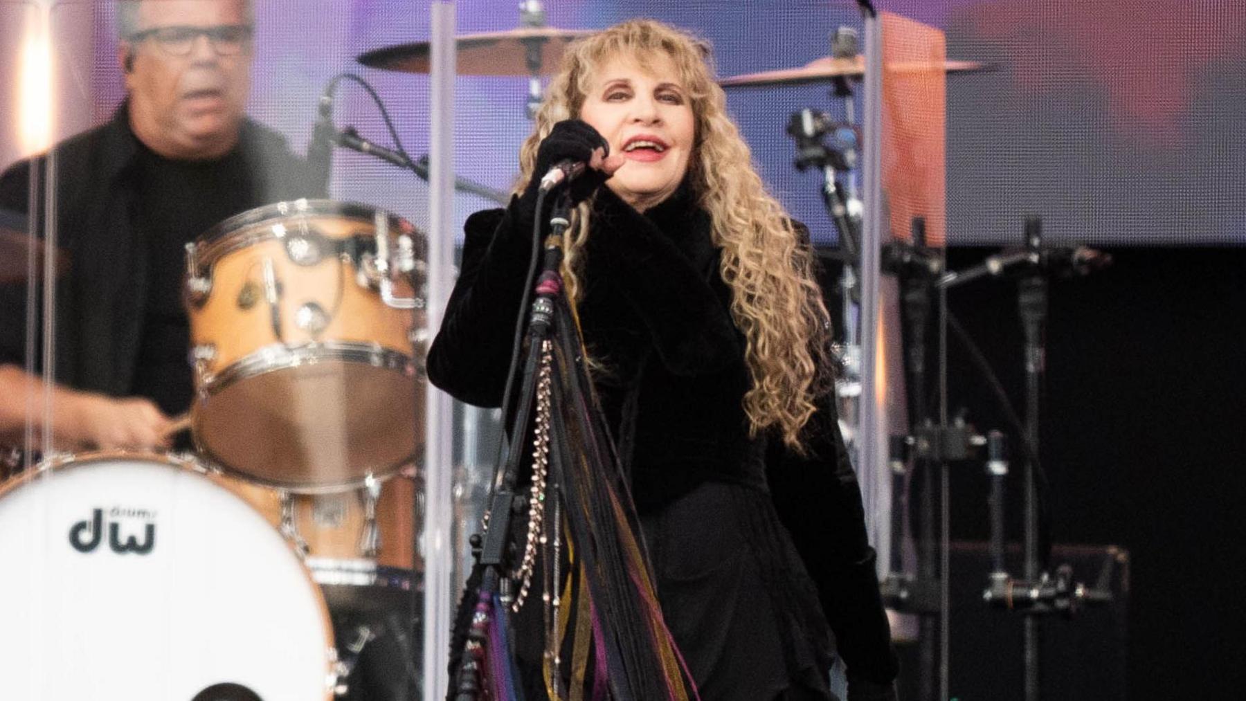 Stevie Nicks on stage holding the microphone with a drummer visible behind her