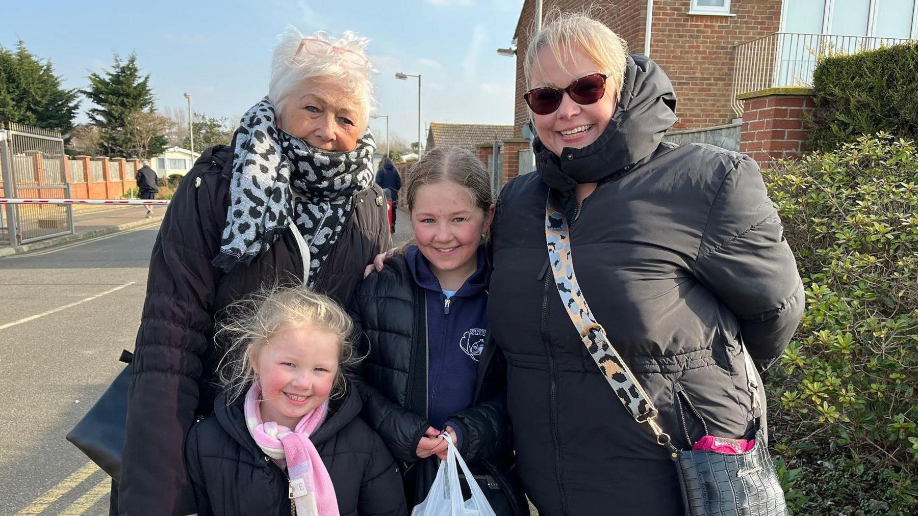 The white family stand bunched together outside of the entrance to the holiday park. They are an older woman with grey hair, a younger woman wearing sunglasses with blonde hair and two female children aged 9 and 5. 