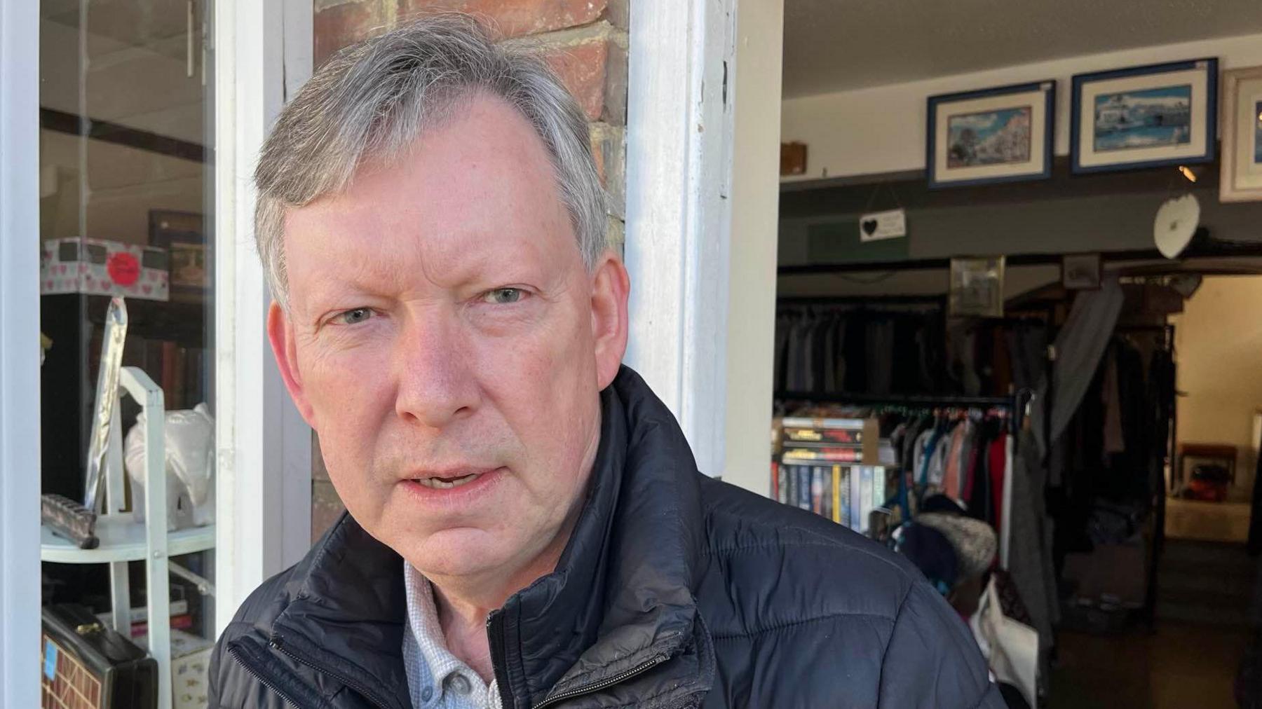 A man with grey hair and a blue jacket stands in front of a charity shop, looking unimpressed.