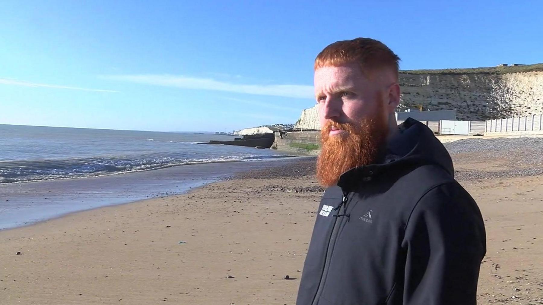 Russ Cook standing on the beach wearing a black jumper. He has a large ginger beard. He is looking slightly away from the camera. 