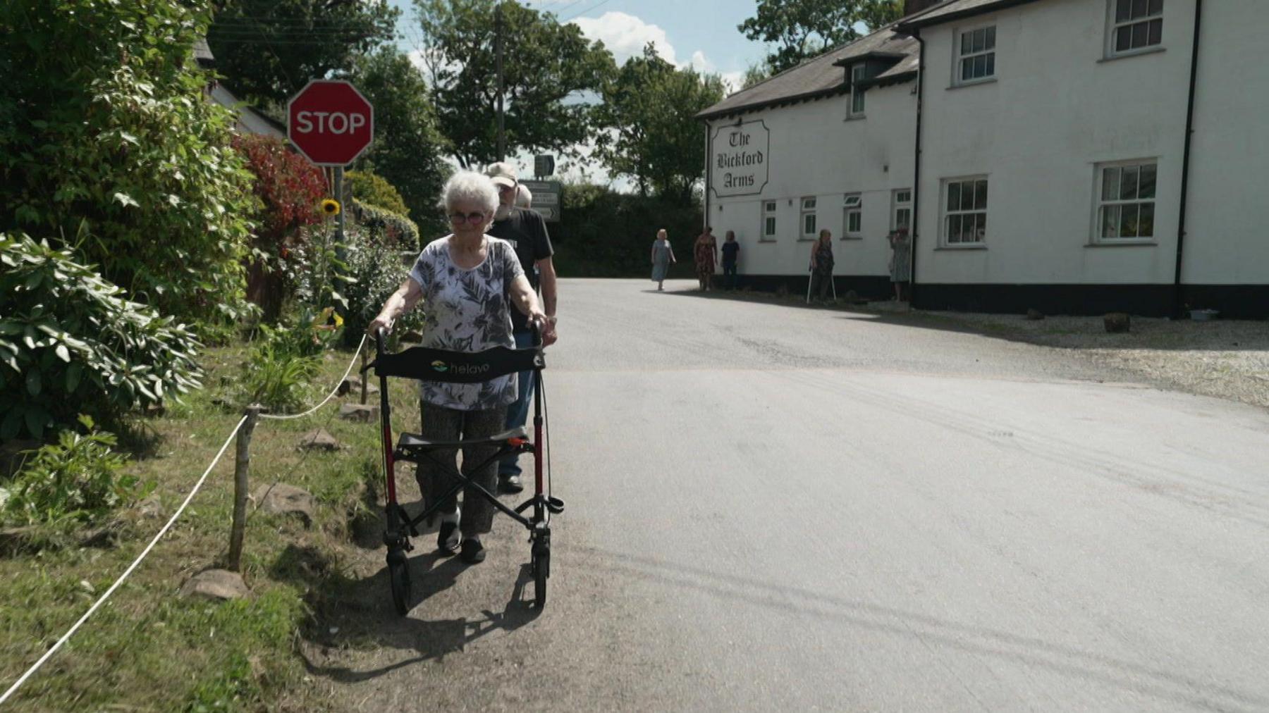 Sylvie Lee who is 89 using a walker to go along the road to her home at Brandis Corner. There people in a column behind her, a red stop sign, and in the background is the local pub and other people walking along the road.