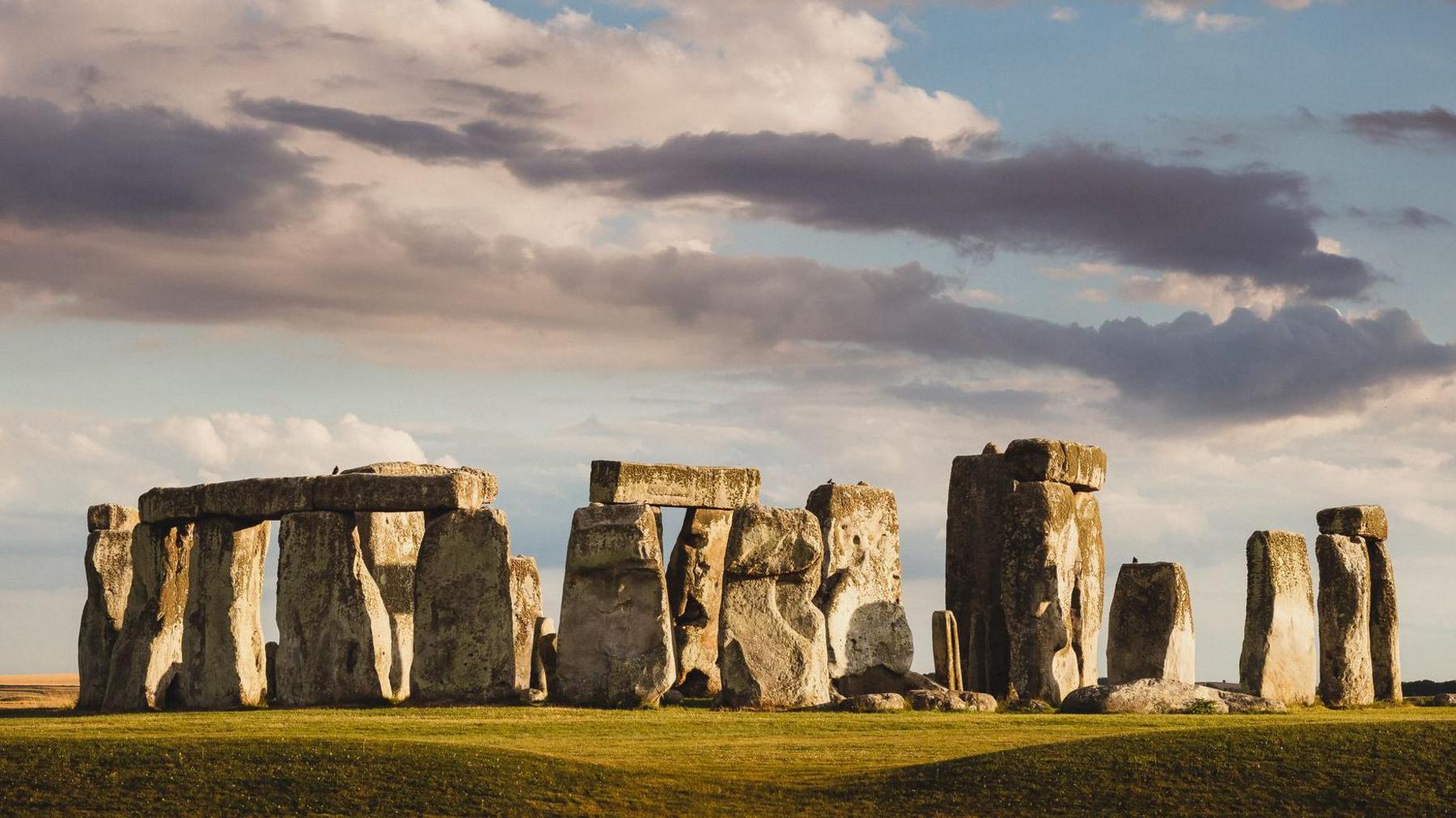 The rocks of Stonehenge stand on green grass. The sky behind them is a bright grey and there are white and dark grey cloud over them. The stones themselves are large. Some have stones lying horizontal across them