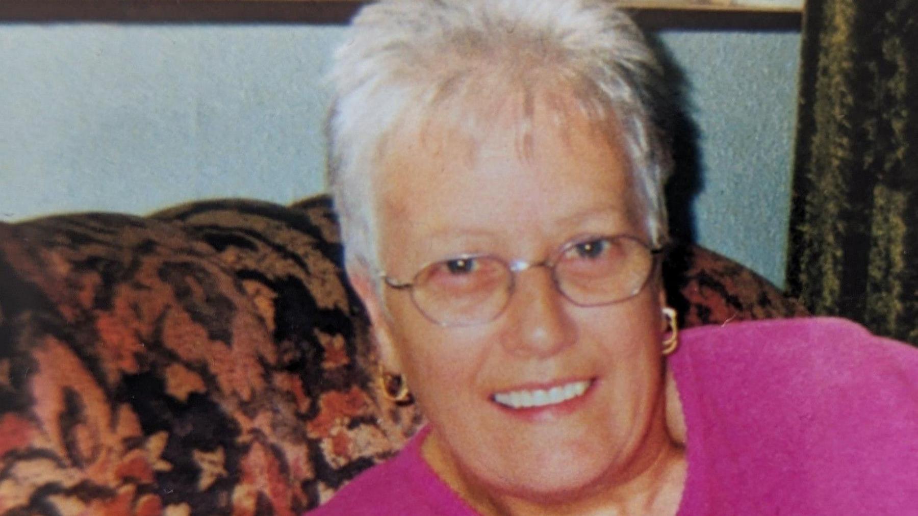 An old photograph - perhaps from the 90s - of an older woman with a short grey pixie cut and oval glasses sitting on an autumnal coloured patterned sofa. She is smiling and is also wearing gold hoops in her ears and a hot pink jumper.