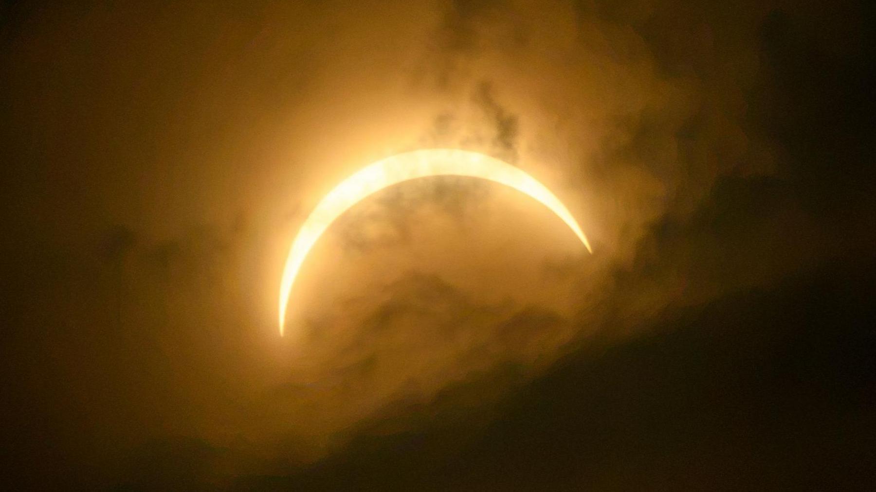 The eclipsed Sun forms a wider, glowing crescent surrounded by warm orange and gold tones. Thick clouds diffuse the sunlight, creating a striking halo effect around the crescent.
