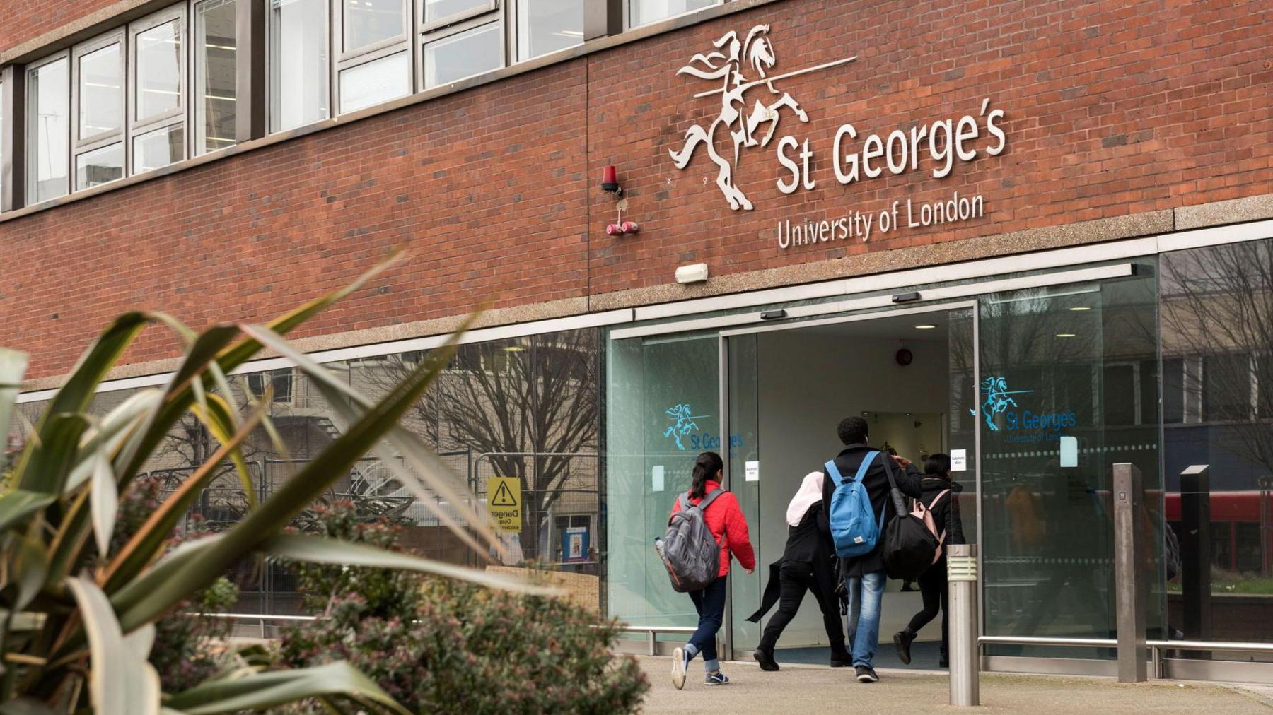 Exterior view of university with students wearing backpacks entering the red brick building through glass doors.