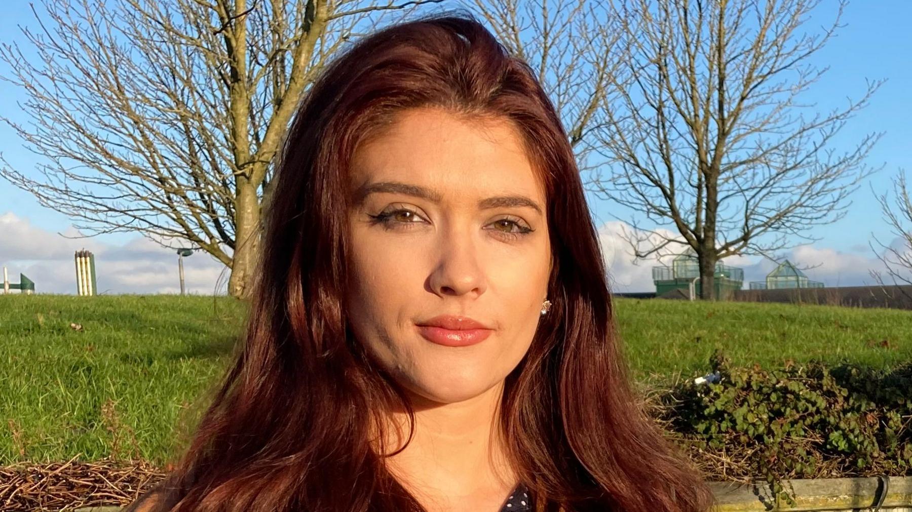 Cara Hunter has long brown hair and is wearing pink lipstick. She is looking straight at the camera. In the background is a green mound of grass with two trees on it - they have no leaves. Behind them is a blue sky.