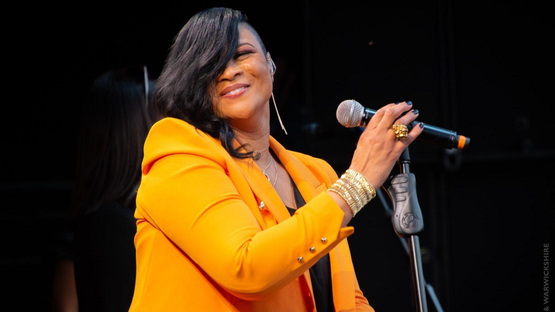 Gabrielle is smiling at the camera while wearing a tangerine-coloured jacket. Her black hair is swept over one side of her face and hangs past her shoulders. She has a large gold ring on her right hand which holds the microphone on its stand