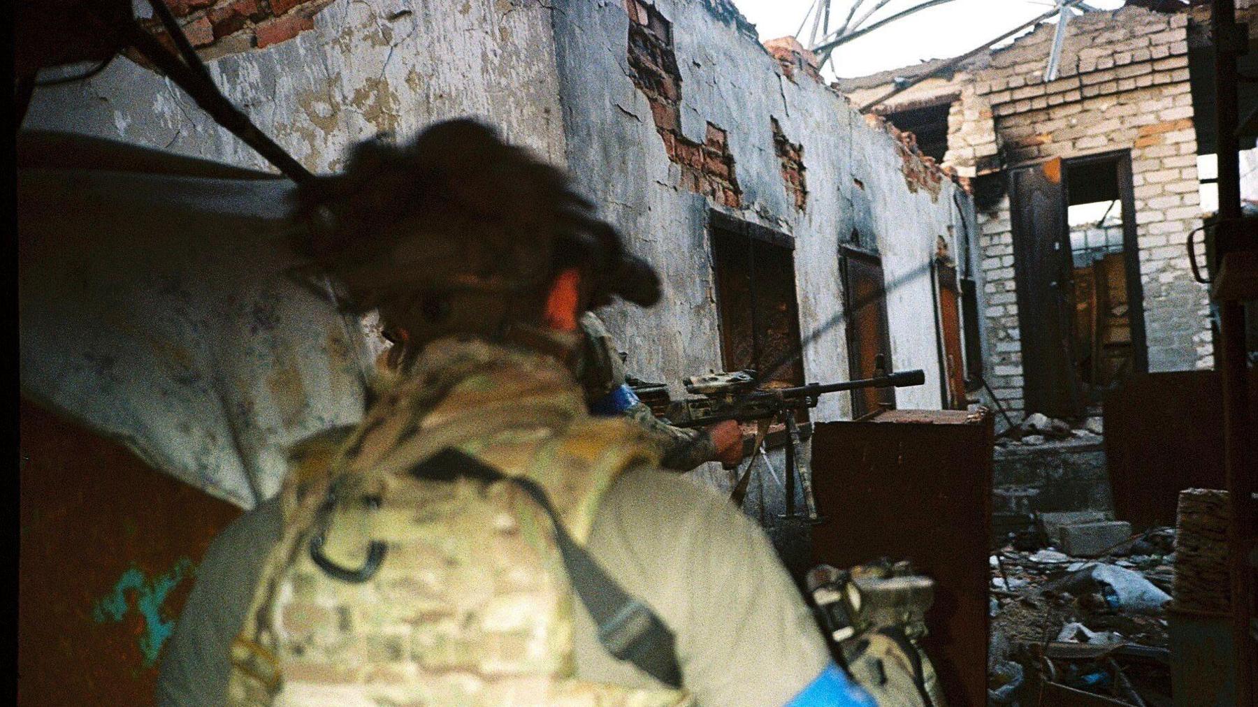 Two unformed Ukrainian soldiers with guns stand outside a heavily bombed building