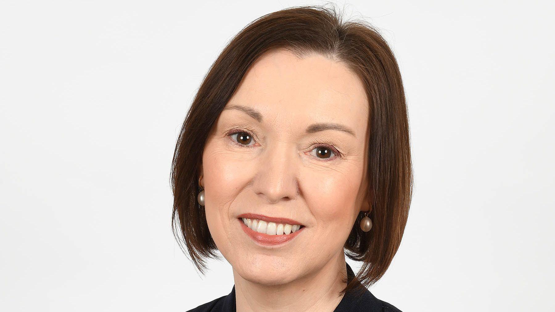 A portrait photo of Jennifer Welsh, who is smiling at the camera and has short brown hair and wearing pearl earrings. She also has black suit jacket on.