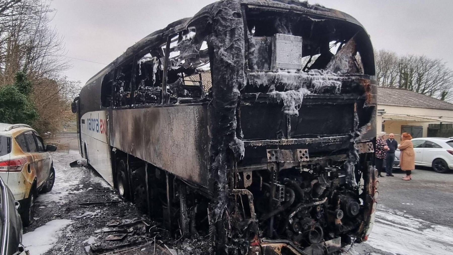 A severely damaged National Express coach. A large section of the vehicle has been left badly burnt and charred with the coach's engine compartment in full view. A white car to its left has also sustained damage from the fire.