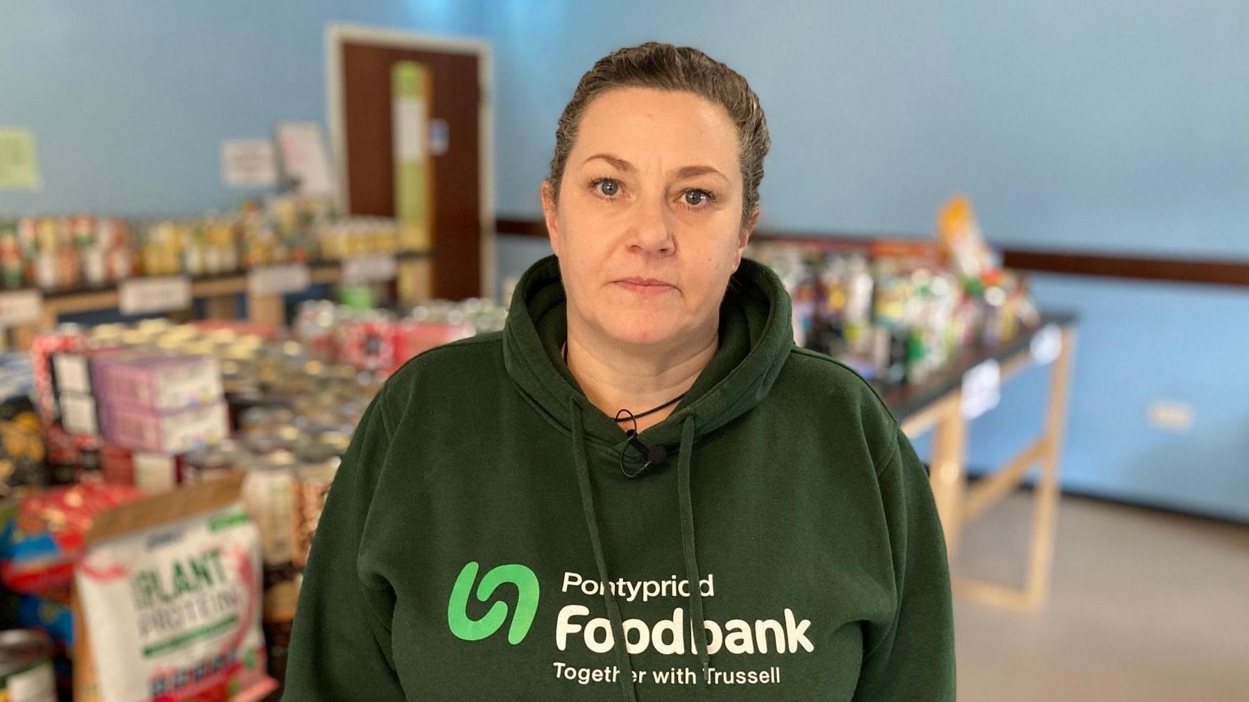 Sharon Elward, cydlynydd banc bwyd Pontypridd yn sefyll o flaen pentyrau o fwyd.  Mae'n gwisgo hwdi gwyrdd sydd a'r geiriau 'Pontypridd Foodbank'.