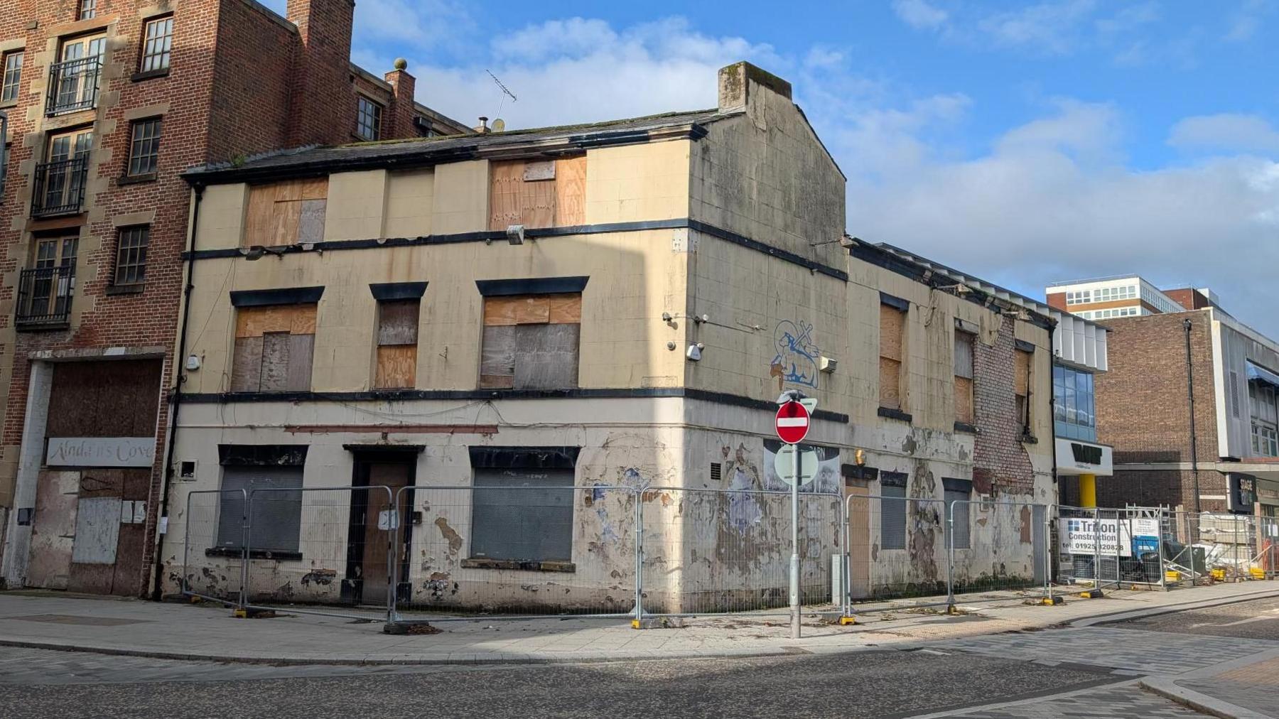 The Tithebarn Pub is an old cream-coloured two-storey building. Its windows are boarded up and the paint work is neglected and cracking. It is surrounded by temporary metal fencing.