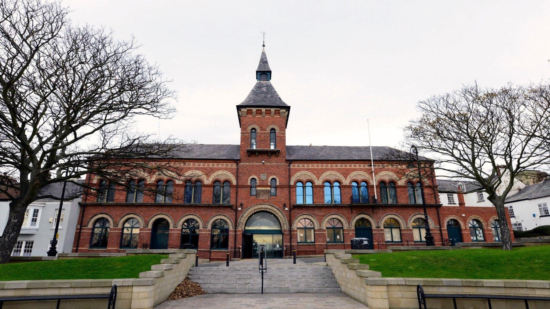 Hartlepool Borough Hall, an Italianate style building in red brick with stone dressings over each of two rows of 11 high-arched windows. In the centre there is a three-stage tower with a wide arched opening on the ground floor and steps leading up to the entrance.