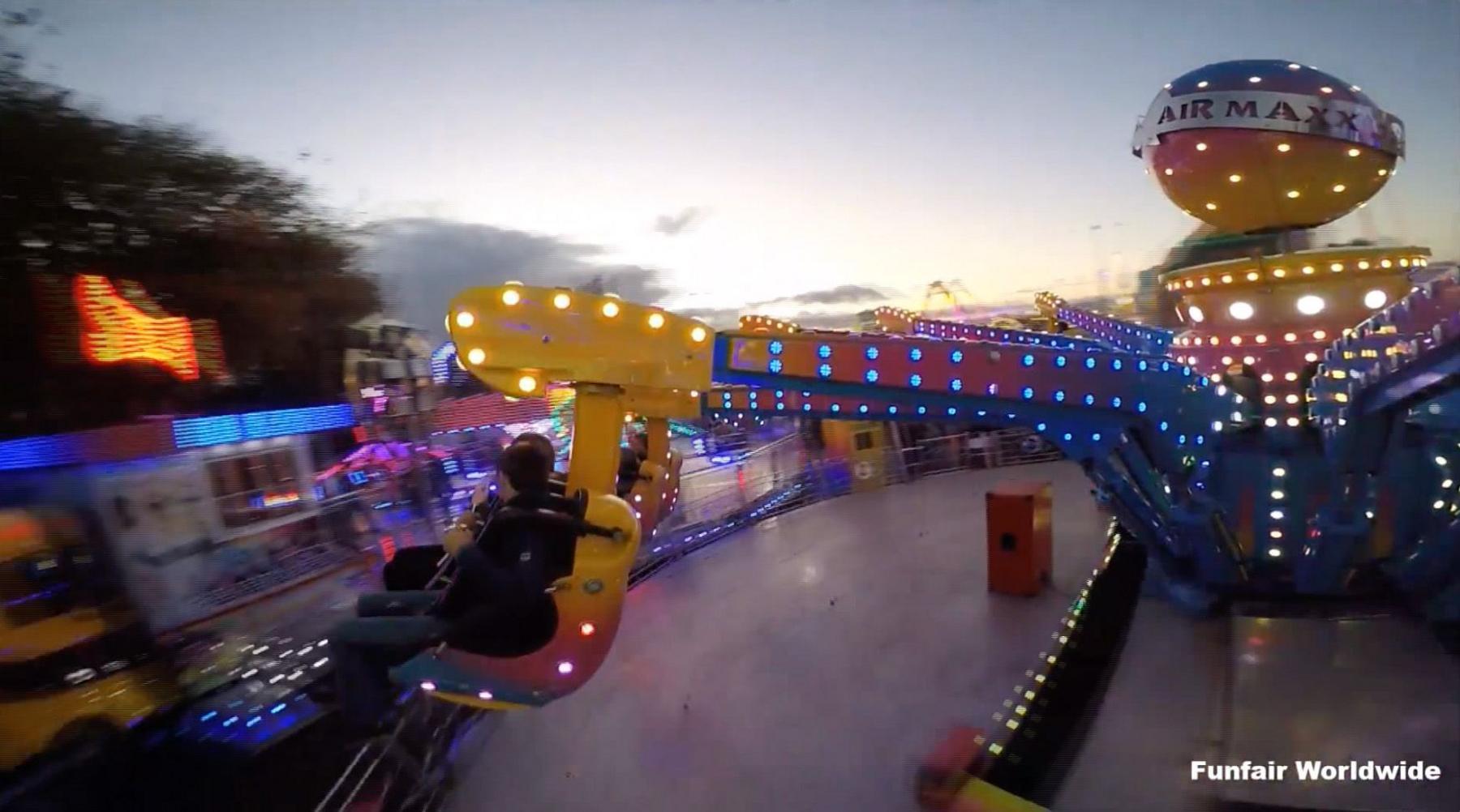 The Air Maxx is captured in motion during dusk. The ride consists of large mechanical arms decorated with vivid lights in blue, yellow, and pink. One of the arms is elevated, holding a seat with two people securely strapped in, facing outward. The background reveals additional illuminated rides and attractions, contributing to the festive atmosphere. On the left side, tall trees frame the scene, while the sky above is partly cloudy, transitioning from daylight to evening.