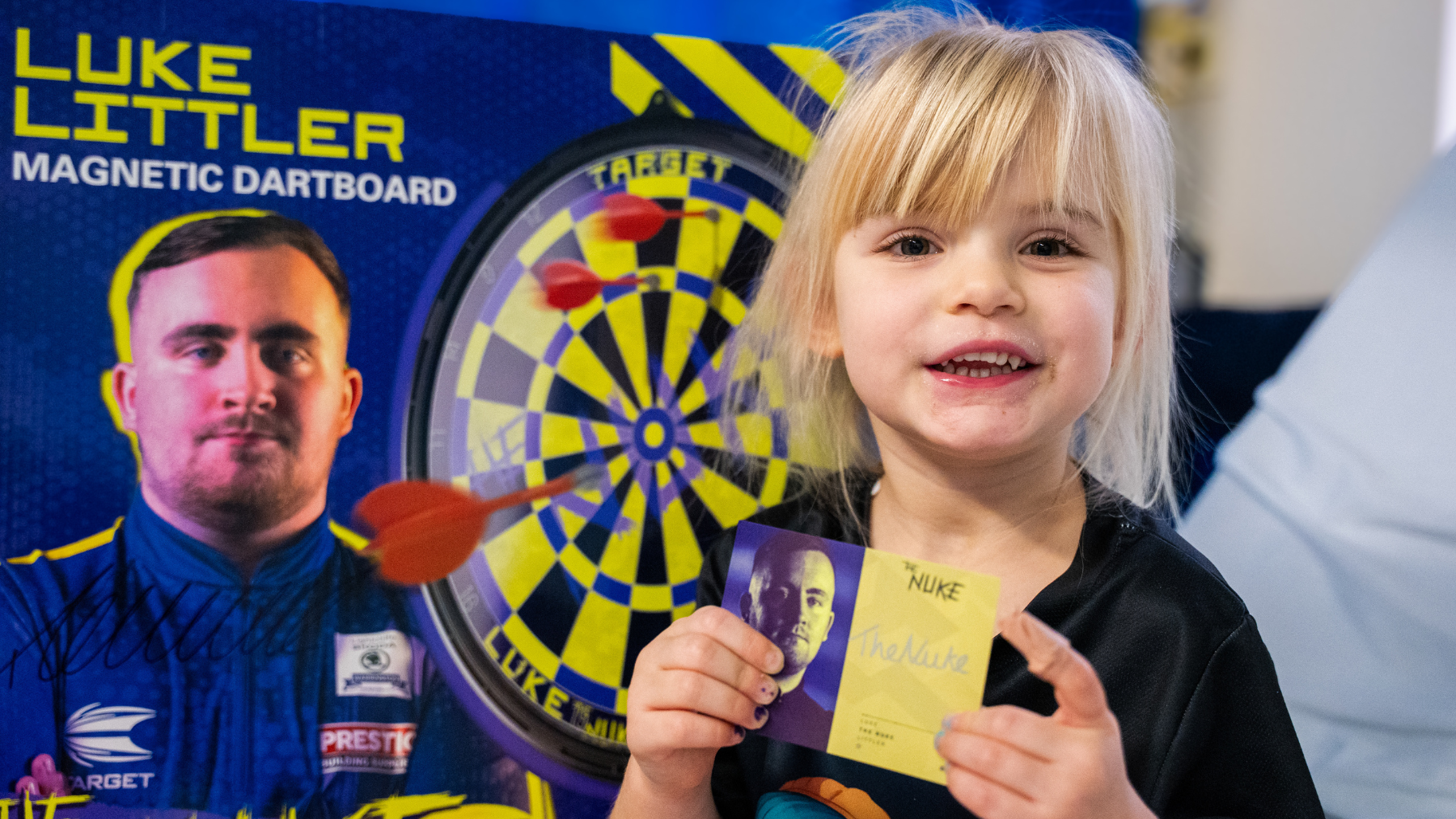 A girl sits in a hospital bed holding a signed card and a magnetic dartboard featuring the name “Luke Littler.” The dartboard design is bright yellow and blue with red darts printed on it. The child wears a dark T-shirt with a colourful cartoon graphic.