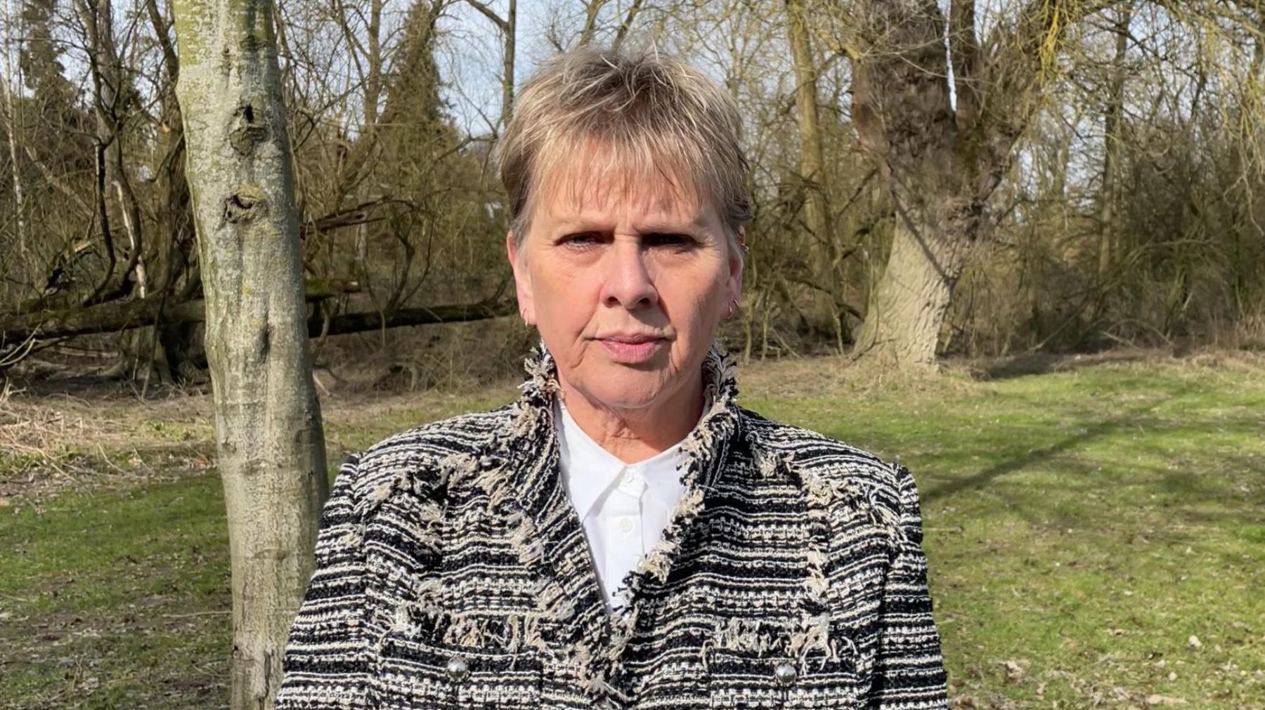 Woman, with blonde hair, wearing a black and white jacket and white shirt, looking expressionless, with a trees and grass behind her.