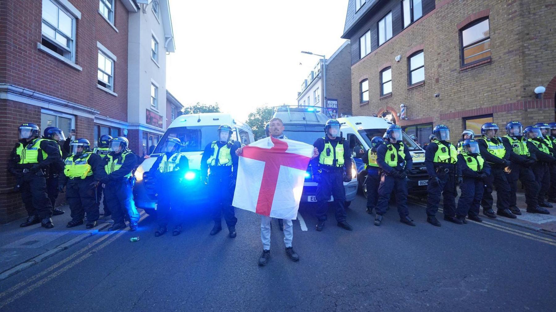 A man holds a St George's flag standing in front of a line of armed police officers with yellow high-vis jackets and face shields. There are flats either side of them and police vans behind them.