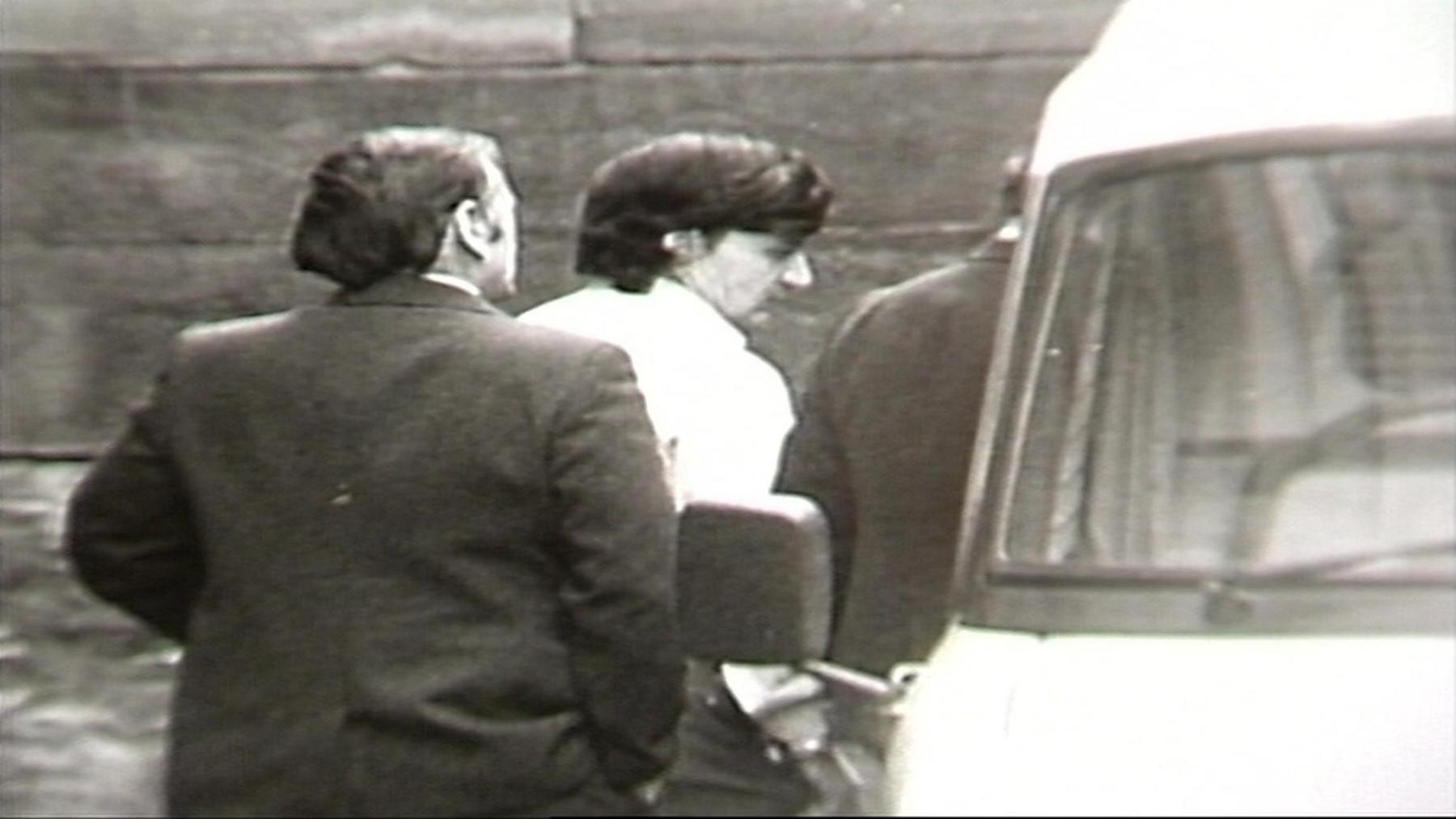 A black and white still image of Peter Sullivan, pictured with thick black hair and a light coloured shirt, being led into a van by two police officers in dark suit jackets