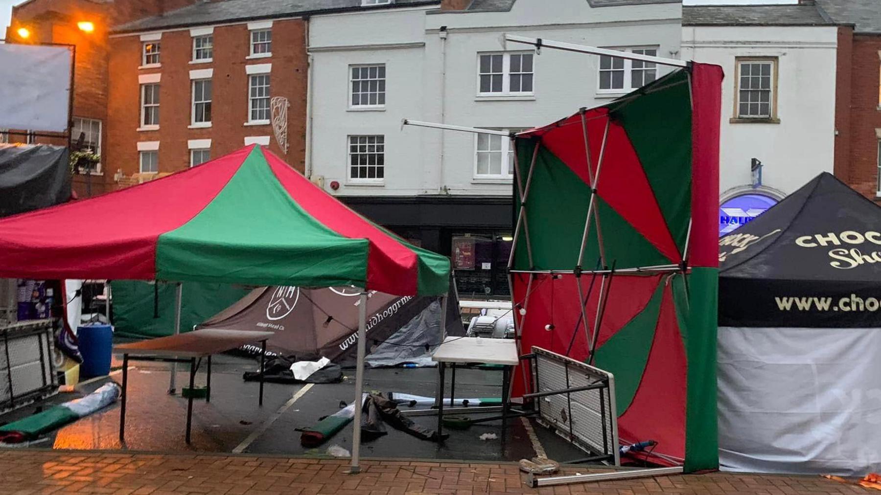 A stall with a red and green roof is blown over on its side. A brown one in the background has collapsed. The ground is littered with bits of metal and fabric.