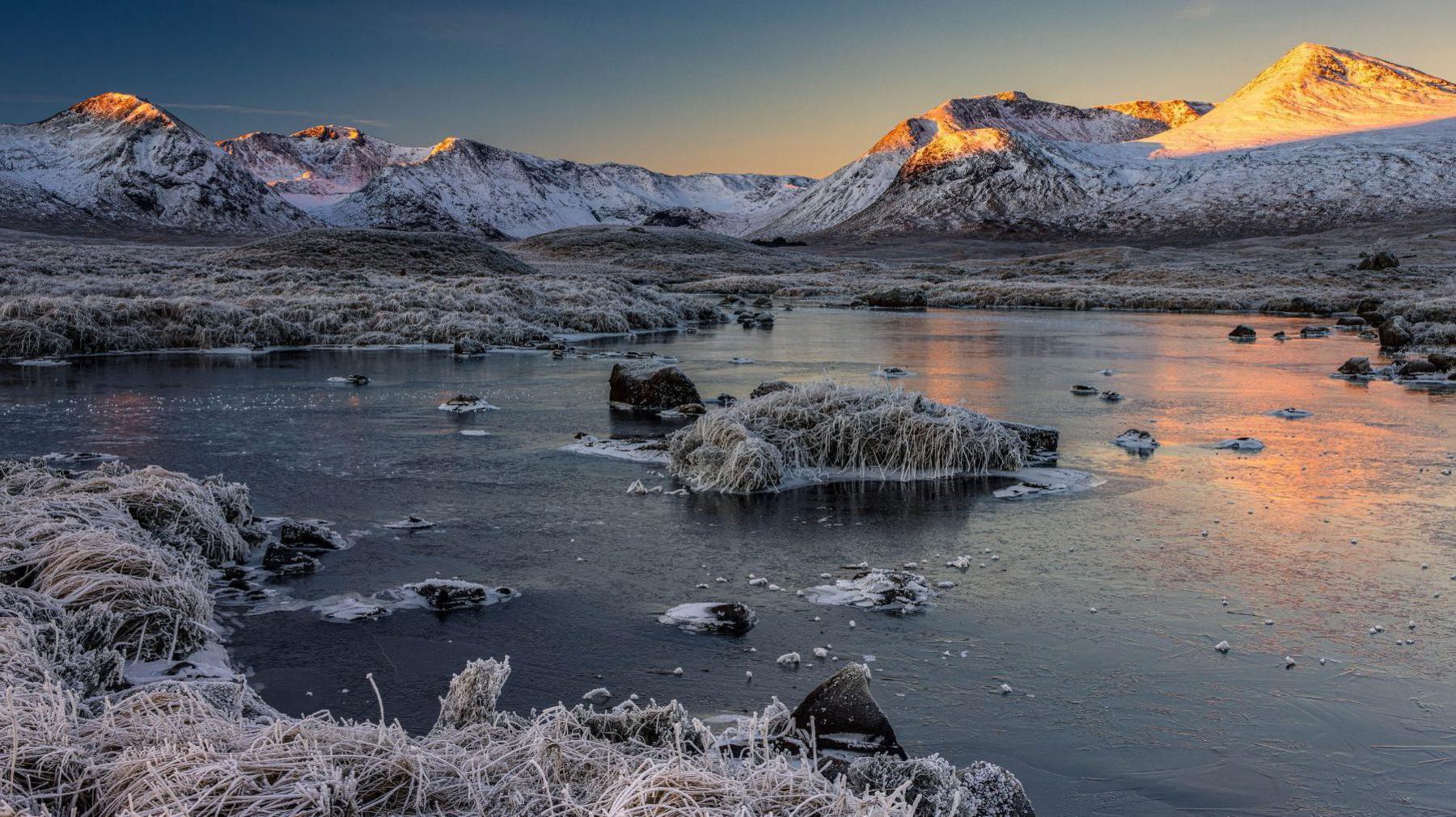 A frozen loch is surrounded by frosted grass with mountains in the background. The water is frozen with a number of rocks poking through the ice. At the centre of the loch is a small island covered in frozen grass. In the background, a snowy mountain range with six peaks covered in snow and dark-coloured land can be seen. Orange light from the morning sun covers two at the far end and the sky is dark blue, fading to light blue and orange.