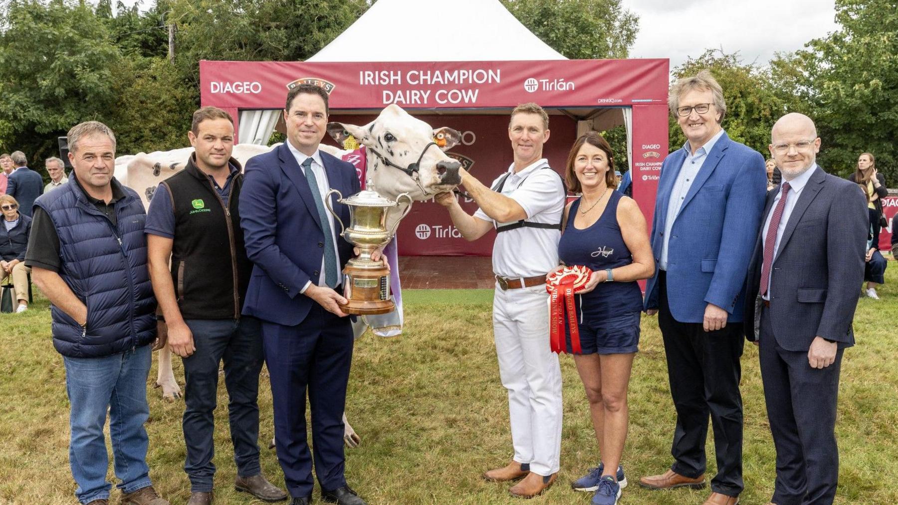 Seven people standing next to the black and white winning cow. From left to right; a man with short grey hair, black t-shirt, navy body warmer, blue jeans and brown boots, then a man with short brown hair, black gilet, navy top and blue jeans with brown boots. Then there is a man in a navy suit with a white shirt amd grey tie holding a gold trophy. Then there is the cow and a man holding it by its' harness, he is dressed in a white top and trousers with a brown belt and brown boots and has blonde hair. Then there is a lady holding a red ribbon who has a navy top, blue denim shorts and blue trainers on, she has short brown hair. There there is a man with grey hair, black glasses and a navy suit jacket with a light shirt and black jeans and brown boots. And finally a bald man with a navy suit, red tie, and white shirt standing at the end. They are standing in front of an 'Irish champion dairy cow' red sign.