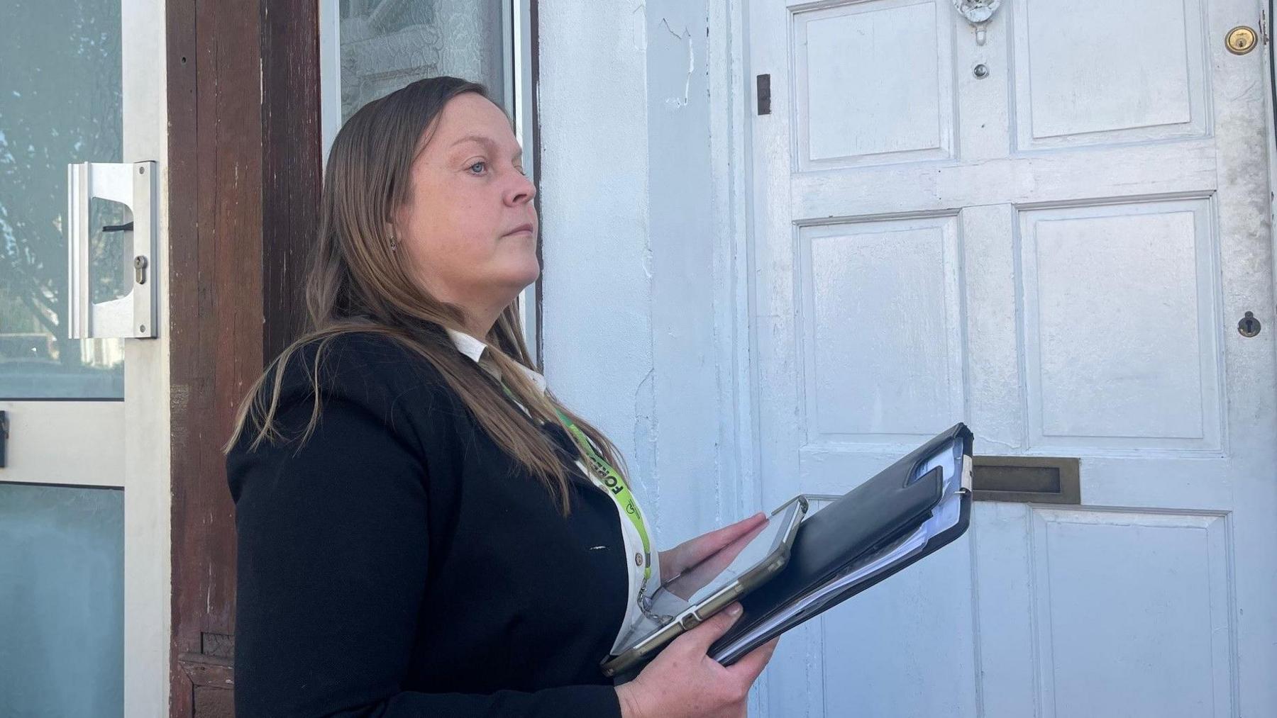A woman with a clipboard and lanyard outside a white-painted wooden door.