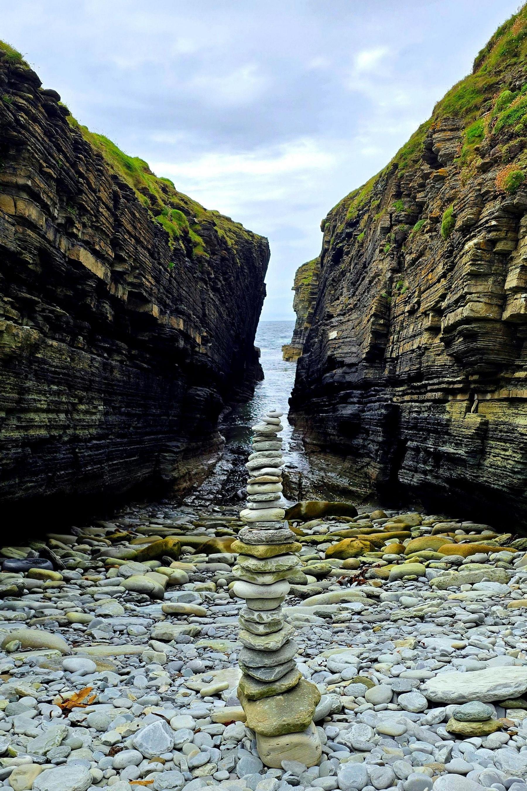 Stones stacked into a tall tower on a rocky shore with the sea and cliffs behind.