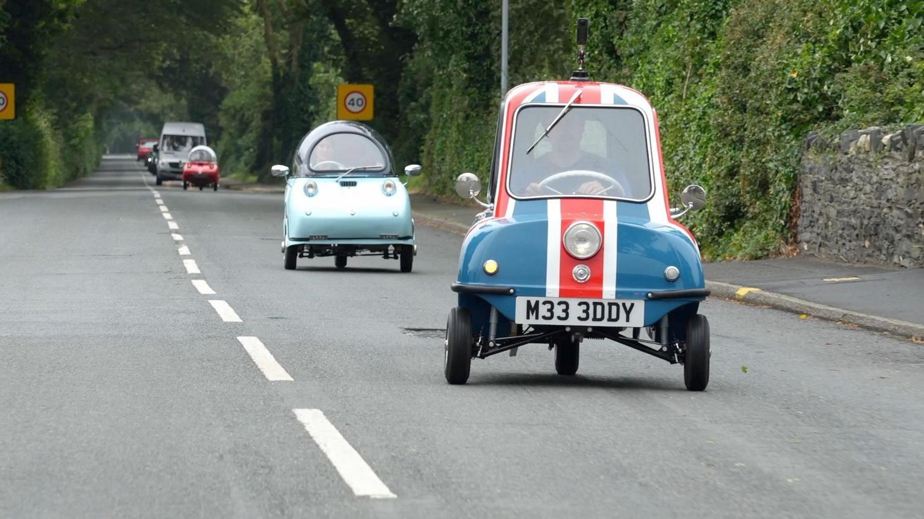 The small Peel P50, with a painted red, white and blue union jack flag drives along a straight road. In the distance, two Tridents, one pale blue, one red follow behind. 