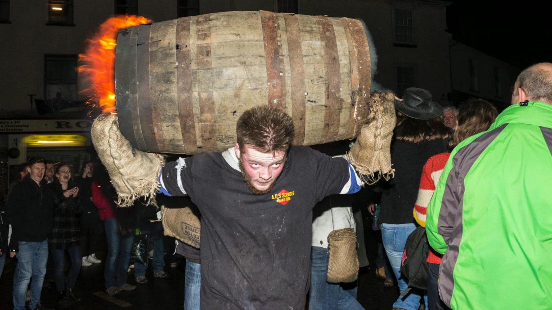 A man runs towards the camera with a barrel on his back. He is wearing dark clothes and has sacking as gloves.