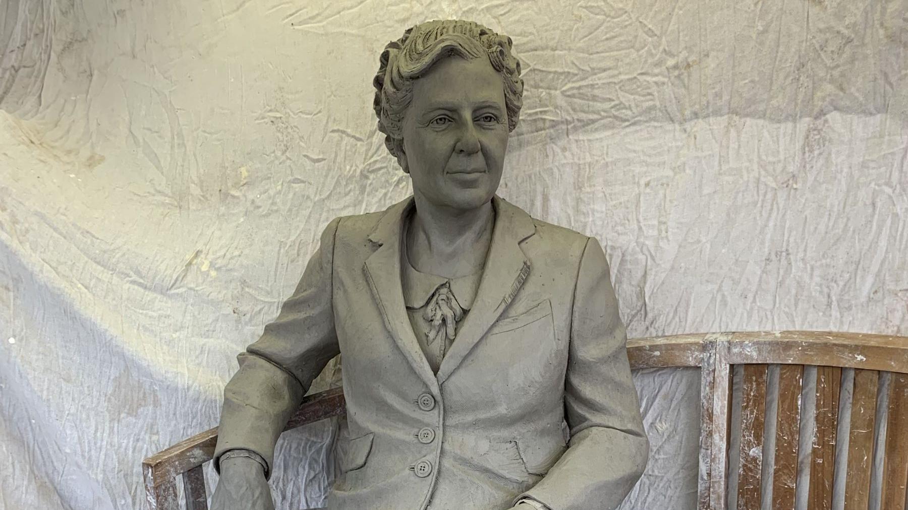 A grey statue of an elderly Agatha Christie in a jacket with short hair. The statue sits on a wooden bench with the arm resting on an arm rest.