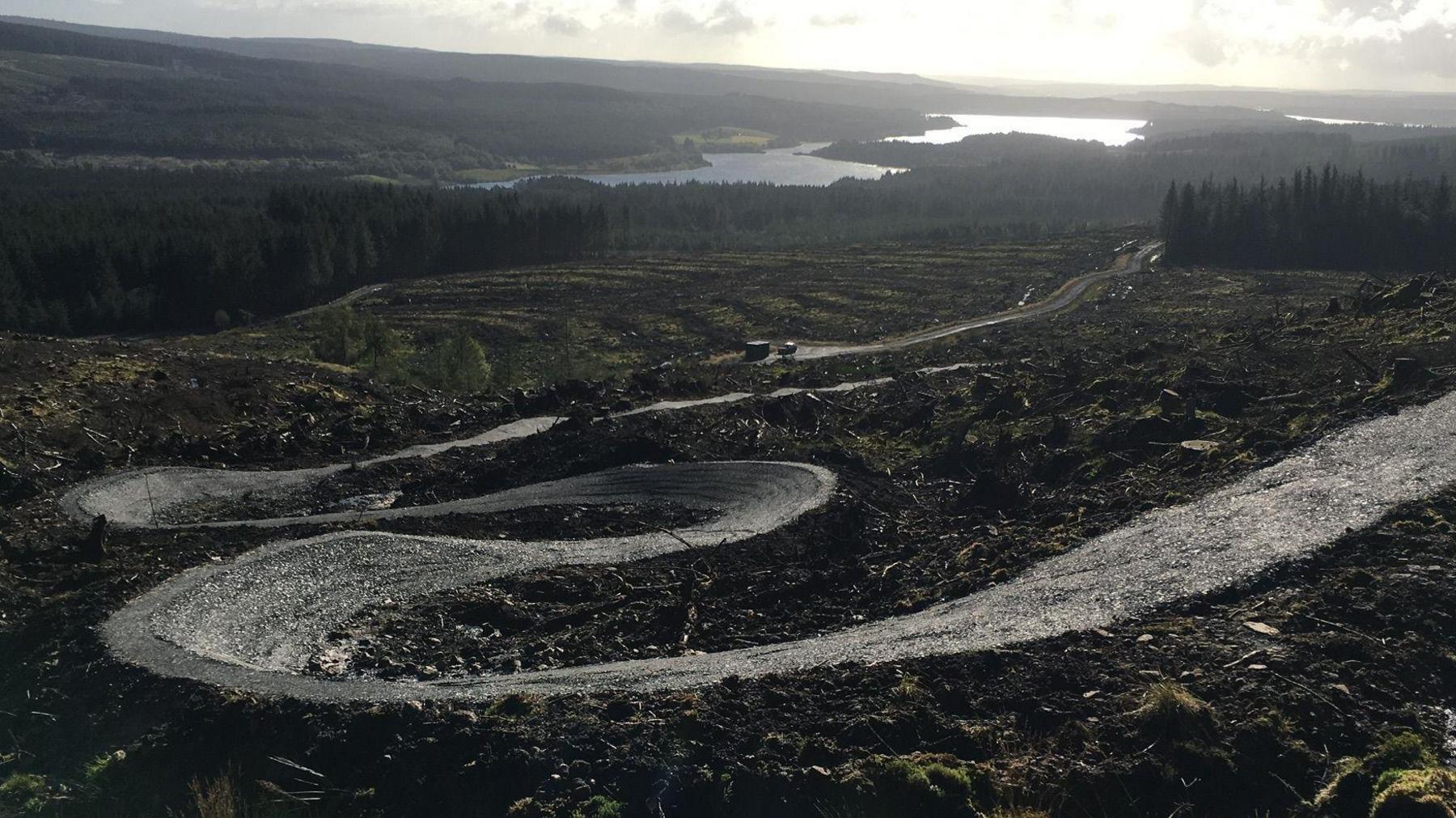 A cycle route with steep sides winds down through an open shrub and moorland landscape with Kielder Reservoir in the distance.