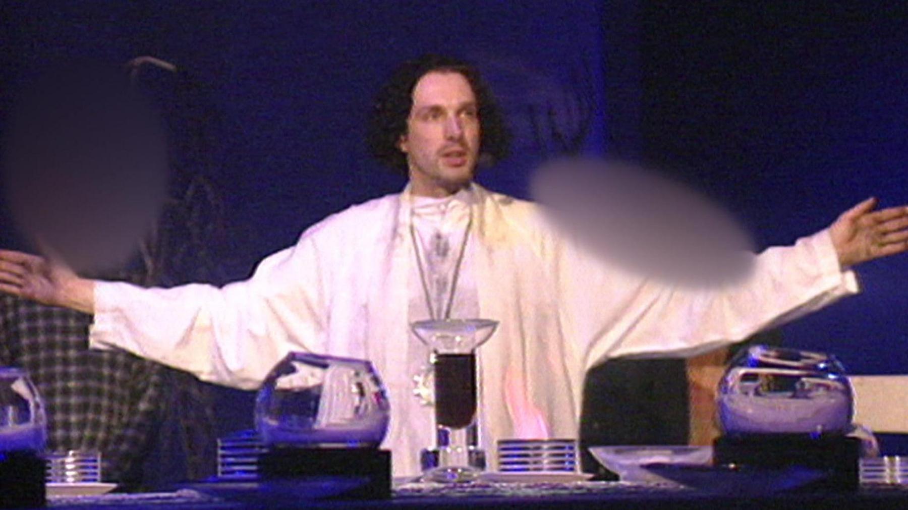 Chris Brain leads a service, wearing white robes and with his arms out wide. The lighting is dim with a blue hue.