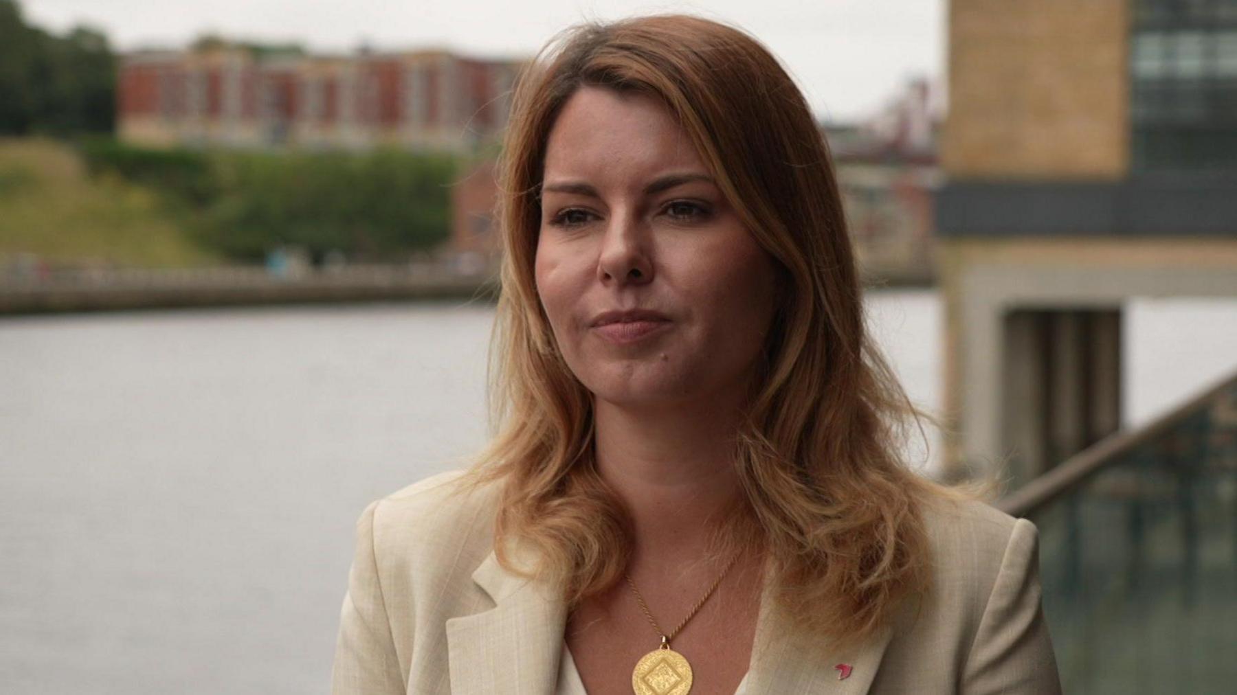Kim McGuinness, who has long blonde hair, stands on the banks of the River Tyne, she is dressed in a beige suit with a gold medallion necklace hanging from her neck.