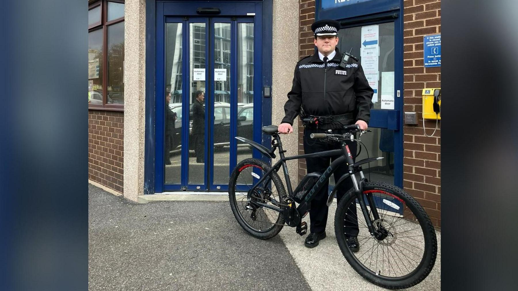 Insp Josh Griffiths stands in front of the entrance to St Helens police station. He is holding a black e-bike.