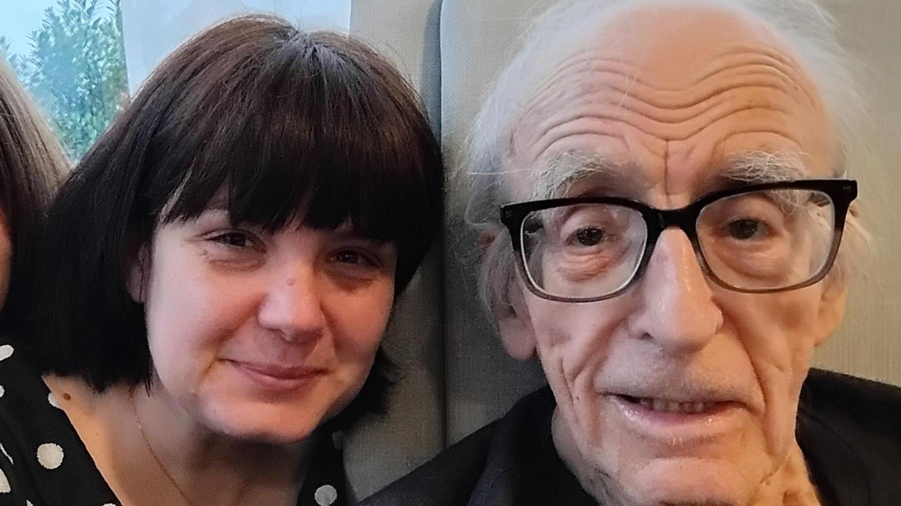 Thre pople pose for a slefie on a grey couch. The person onnthe far right is an elderly gentleman with thin grey hair and glasses. The women in the middle has on a black top with white Polka dots, she has brown hair and a fringe. The boy on the far left of the three has long brown hair. Half his face is cropped out of the photo.